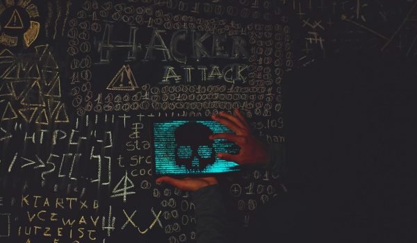 A hacker in a black hoodie using a tablet displaying a skull, surrounded by chalk symbols and 'Hacker Attack' text.